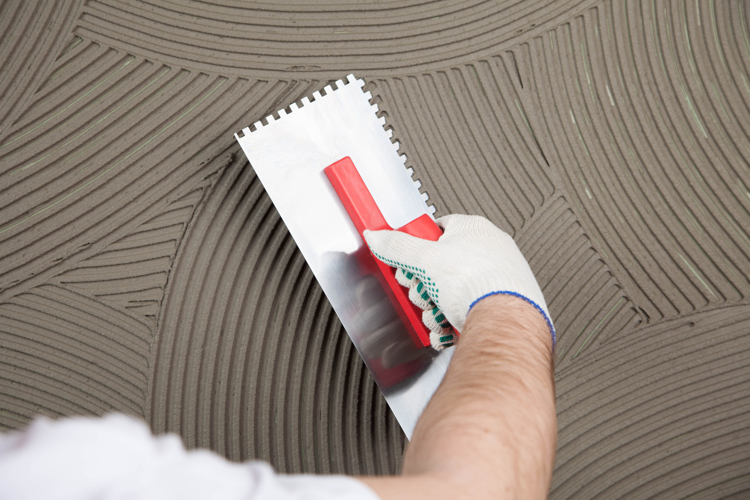 A hand holds a notched trowel, applying wet mortar to a surface, creating textured lines. This showcases plastering techniques for construction projects.